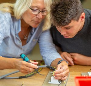 Eine ältere Frau und ein junger Mann arbeiten gemeinsam an einem technischen Projekt, während sie ein Lötkolben benutzen. Sie konzentrieren sich auf die Details der Arbeit und zeigen eine kooperative Lernumgebung.