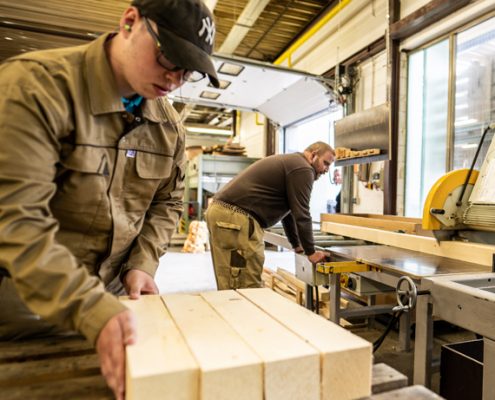 Handwerker in einer Holzwerkstatt, der präzise Holzstücke bearbeitet, während ein Kollege im Hintergrund arbeitet. Helle, gut beleuchtete Umgebung mit professioneller Ausrüstung.