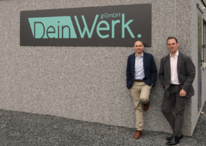 Zwei Männer stehen an einer Wand mit dem Schriftzug "DeinWerk." im Hintergrund. Die Szene vermittelt eine professionelle Atmosphäre, ideal für Unternehmenspräsentationen oder Networking-Events.