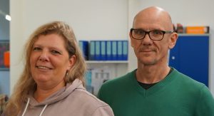 Zwei Personen stehen vor einer Tür. Eine Frau mit Hoodie lächelt. Ein Mann mit Brille schaut nach vorne.