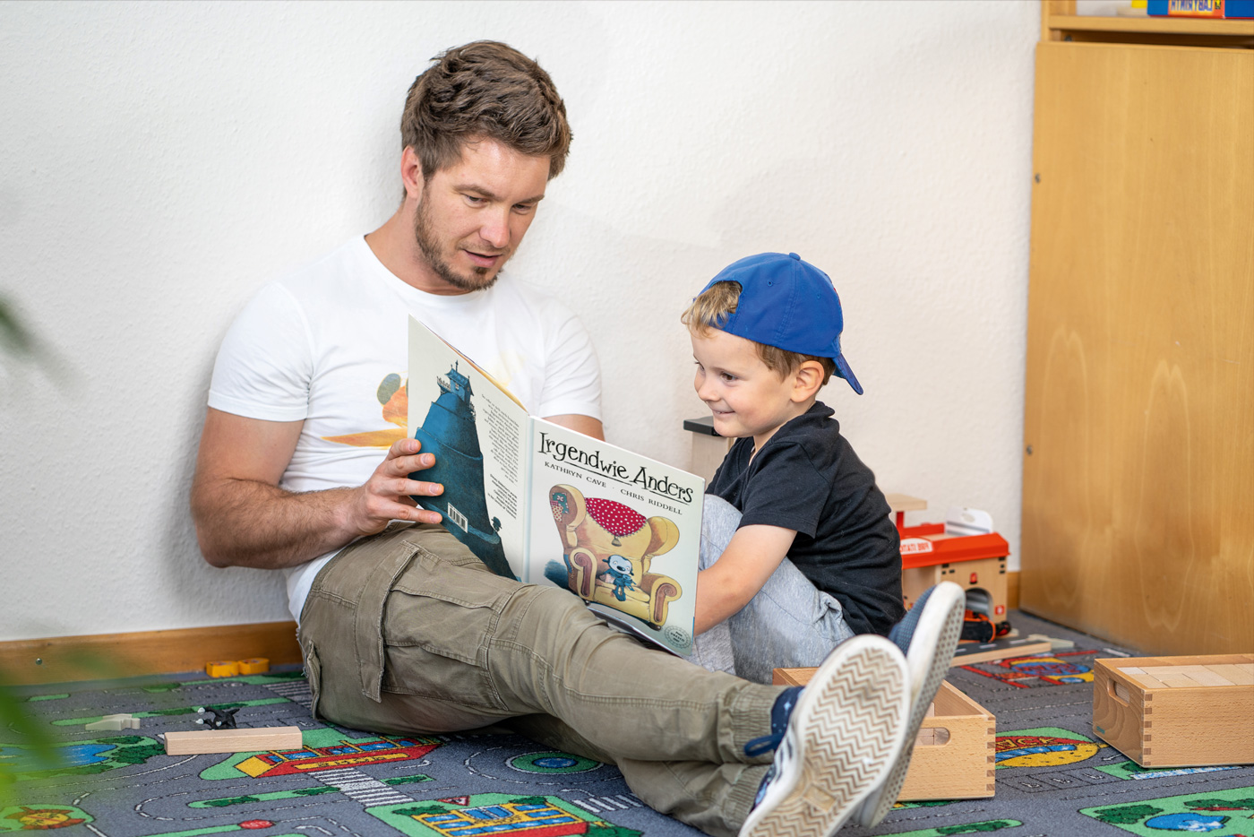 Vater und Sohn lesen gemeinsam ein Buch auf dem Boden, umgeben von Spielzeug. Der Vater trägt ein T-Shirt und der Junge eine Kappe, beide wirken konzentriert und glücklich.