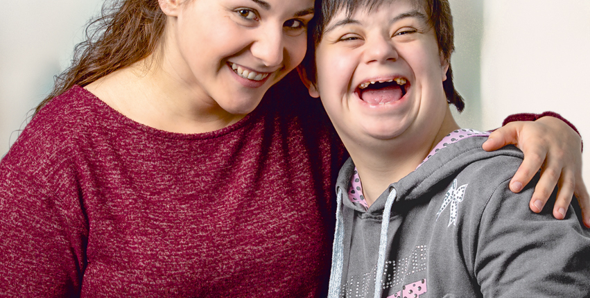 Zwei lächelnde Frauen umarmen sich herzlich, eine von ihnen hat Down-Syndrom. Die Szene strahlt Freude und Freundschaft aus.