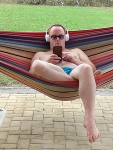 Mann liegt in einer Hängematte. Er trägt keine Oberteile und hört Musik mit Kopfhörern.