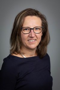 Porträt einer Frau mit Brille und mittellangen Haaren, die in einem professionellen Umfeld lächelt.