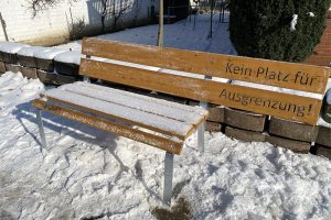 Holzbank im Schnee mit einer schneebedeckten Sitzfläche, umgeben von winterlicher Landschaft.