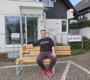 Person sitzt auf einer Holzbank vor einem modernen Gebäude, umgeben von einer ruhigen Umgebung. Die Szene vermittelt Entspannung und lädt zum Verweilen ein.