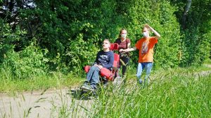 Ein Junge sitzt im Rollstuhl. Zwei andere Kinder helfen ihm auf einem Weg.