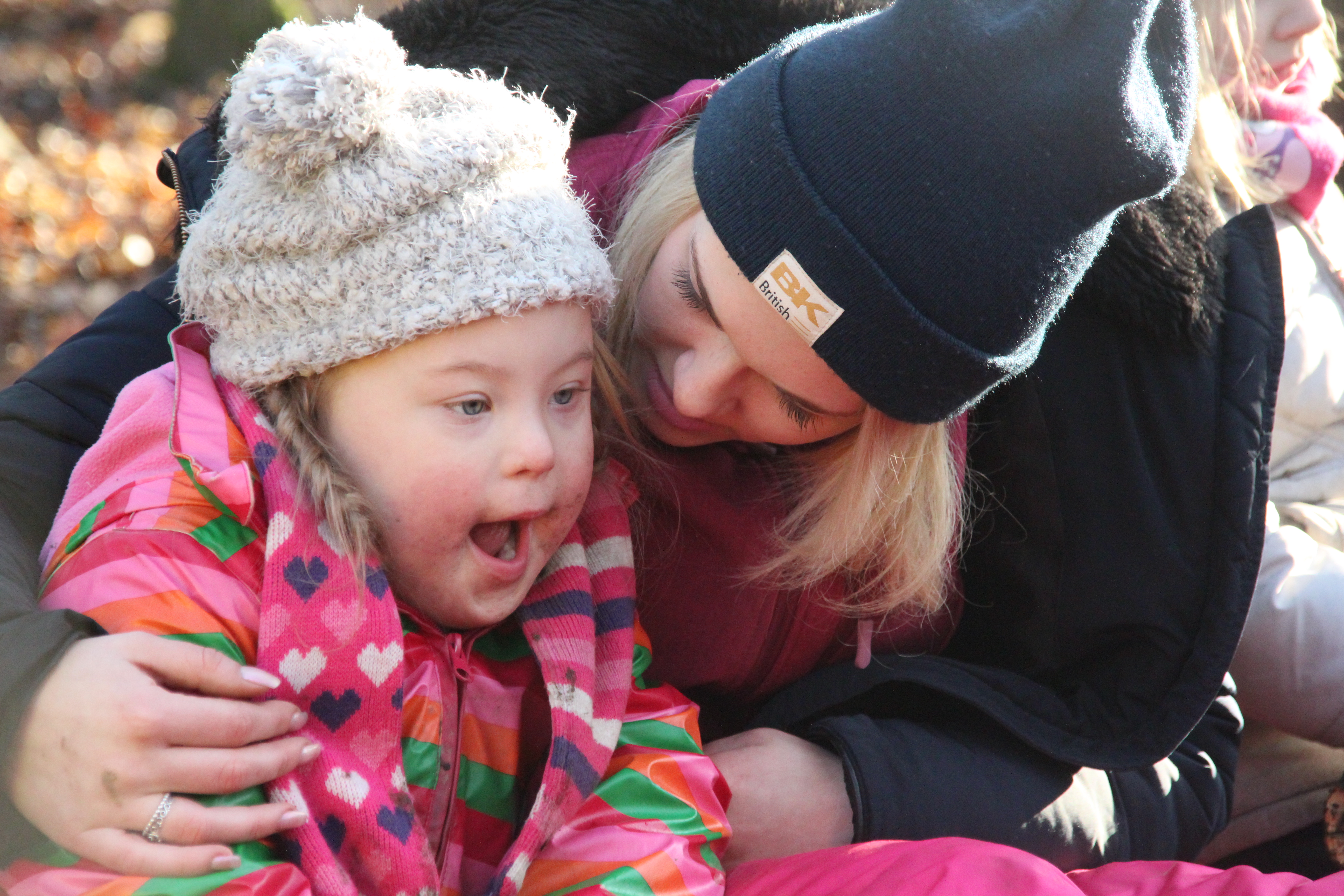Lächelnde Mutter und Tochter genießen einen sonnigen Tag im Freien, während das Kind in einem bunten Mantel und einer Mütze strahlt.