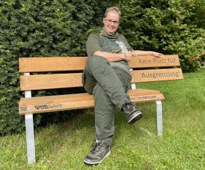 Mann sitzt entspannt auf einer Holzbank im Garten, umgeben von grünen Pflanzen, trägt lässige Kleidung und schaut in die Kamera.