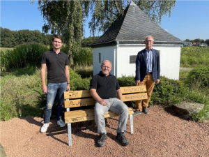 Drei Männer stehen und sitzen auf einer Bank in einem Garten, im Hintergrund ein kleiner Pavillon und Bäume.