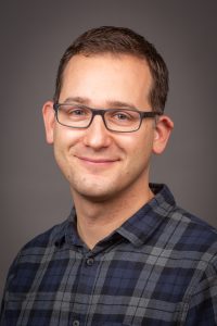 Porträt eines lächelnden Mannes mit Brille, der ein kariertes Hemd trägt. Der Hintergrund ist neutral und sorgt für einen professionellen Eindruck. Ideal für Business-Profile oder Teamvorstellungen.