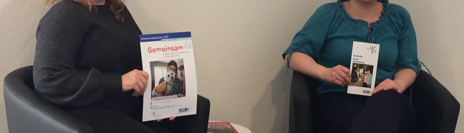Zwei Frauen sitzen in einem modernen Wartezimmer und halten Informationsbroschüren in der Hand. Im Hintergrund sind bunte Bilder an der Wand zu sehen.