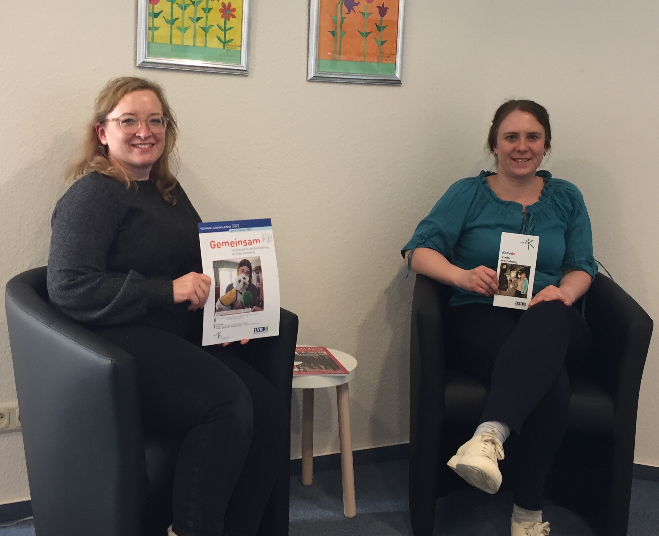 Zwei Frauen sitzen in einem modernen Wartezimmer und halten Informationsbroschüren in der Hand. Im Hintergrund sind bunte Bilder an der Wand zu sehen.