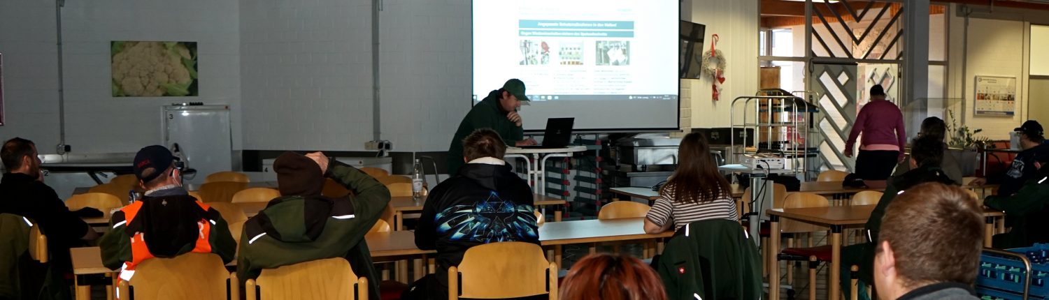 Ein Referent präsentiert eine digitale Präsentation vor einer Gruppe von Zuhörern in einem Seminarraum mit Holzbalkendecke und Tischen.