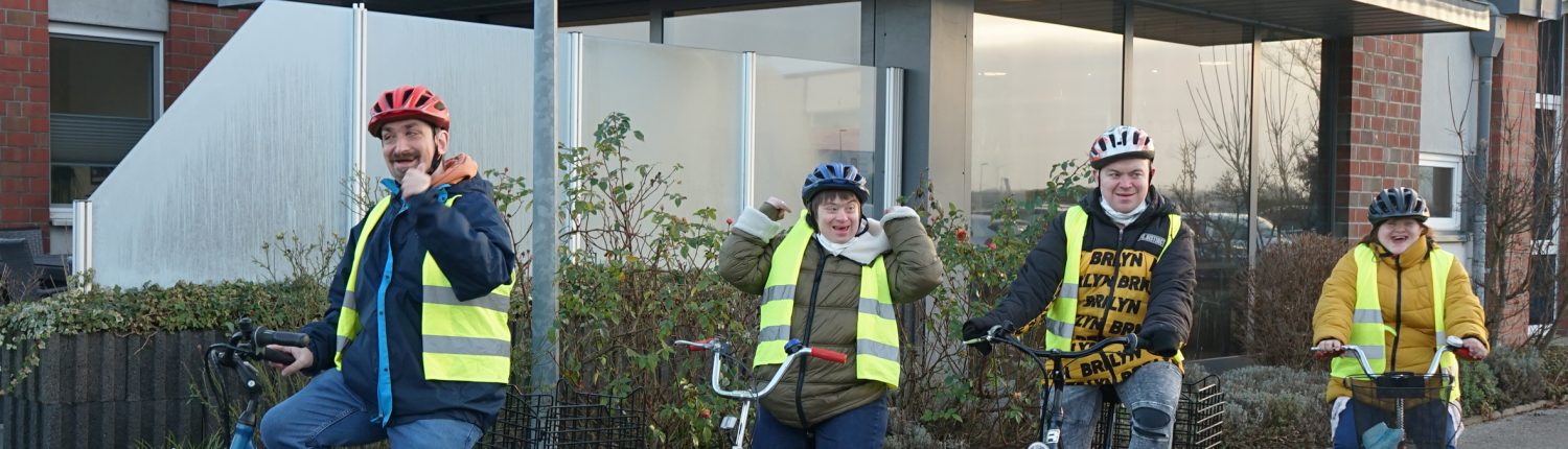 Fünf Personen fahren mit Fahrrädern vor einem Gebäude. Alle tragen eine Warnweste und einen Helm.