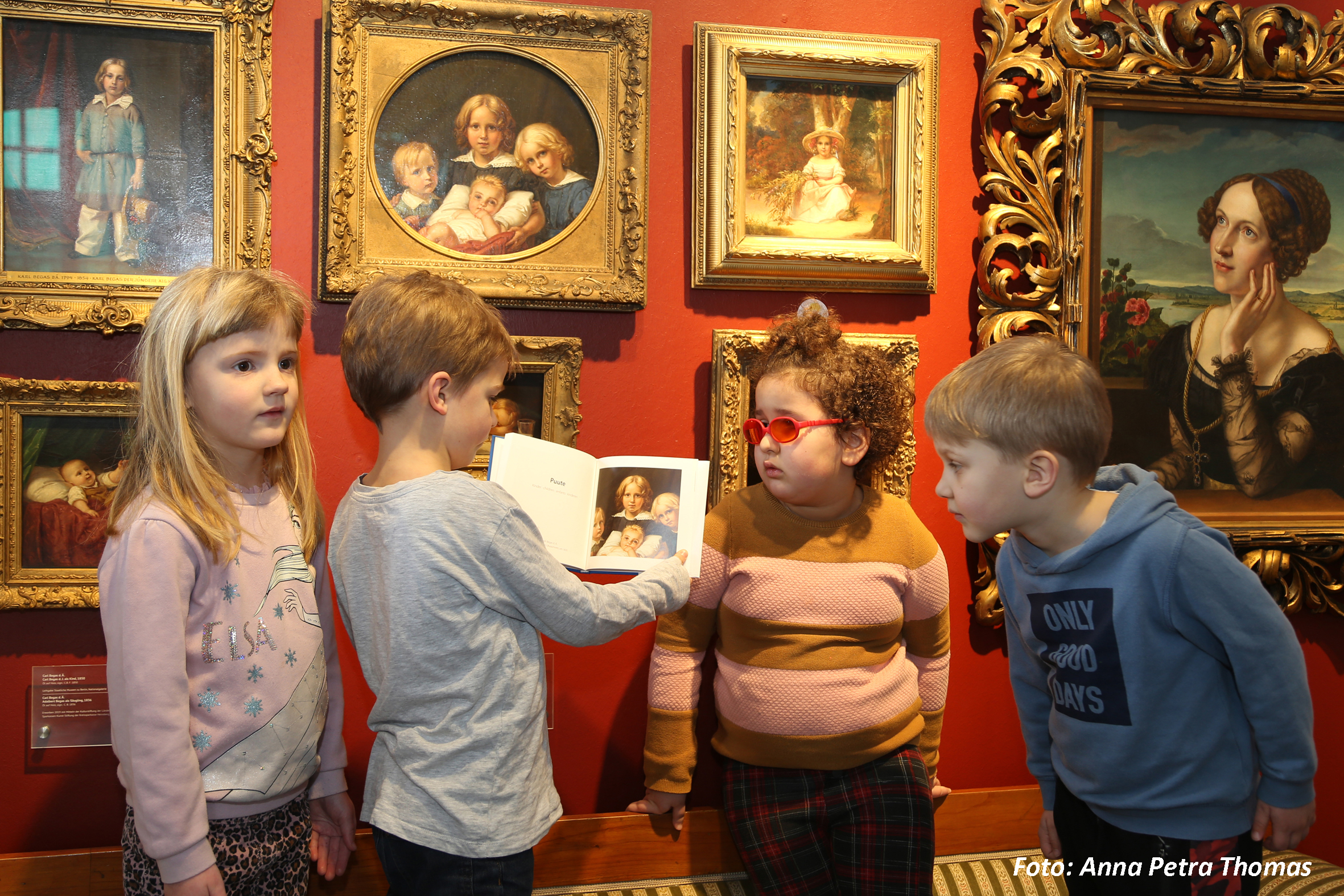 Kinder stehen vor einem Gemälde. Ein Junge hält ein Buch. Sie schauen alle auf das Bild.