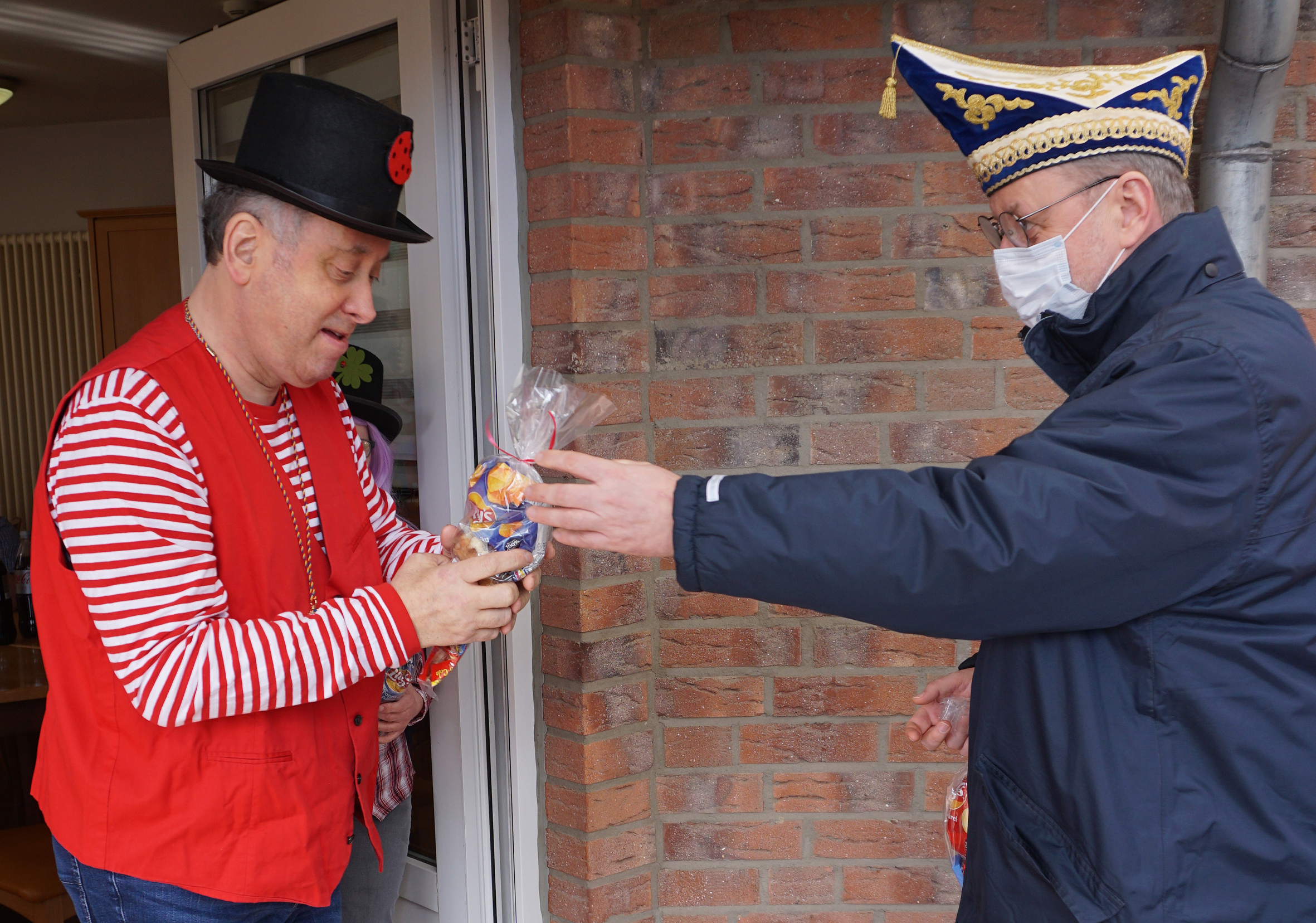 Zwei Männer in Festtagskleidung stehen vor einem Gebäude. Der Mann links trägt einen roten Anzug und einen Zylinder, während der Mann rechts eine Mütze und eine Maske trägt. Sie übergeben sich ein Geschenk oder eine Urkunde, was auf eine Feier oder eine besondere Veranstaltung hindeutet.