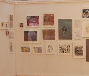 Wand mit verschiedenen Kunstwerken in einer Galerie, darunter Fotografien und Drucke, die zeitgenössische Themen darstellen.