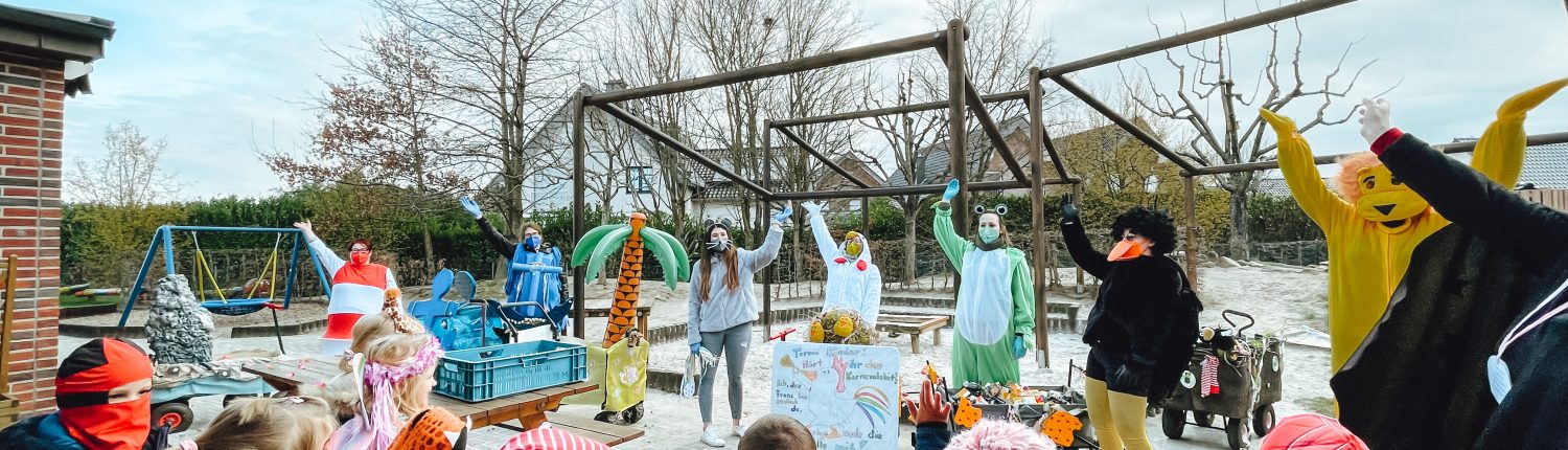 Kinder feiern draußen mit einem Maskottchen. Manche tragen Kostüme und Hüte. Es gibt einen kleinen See und einen Spielplatz im Hintergrund.