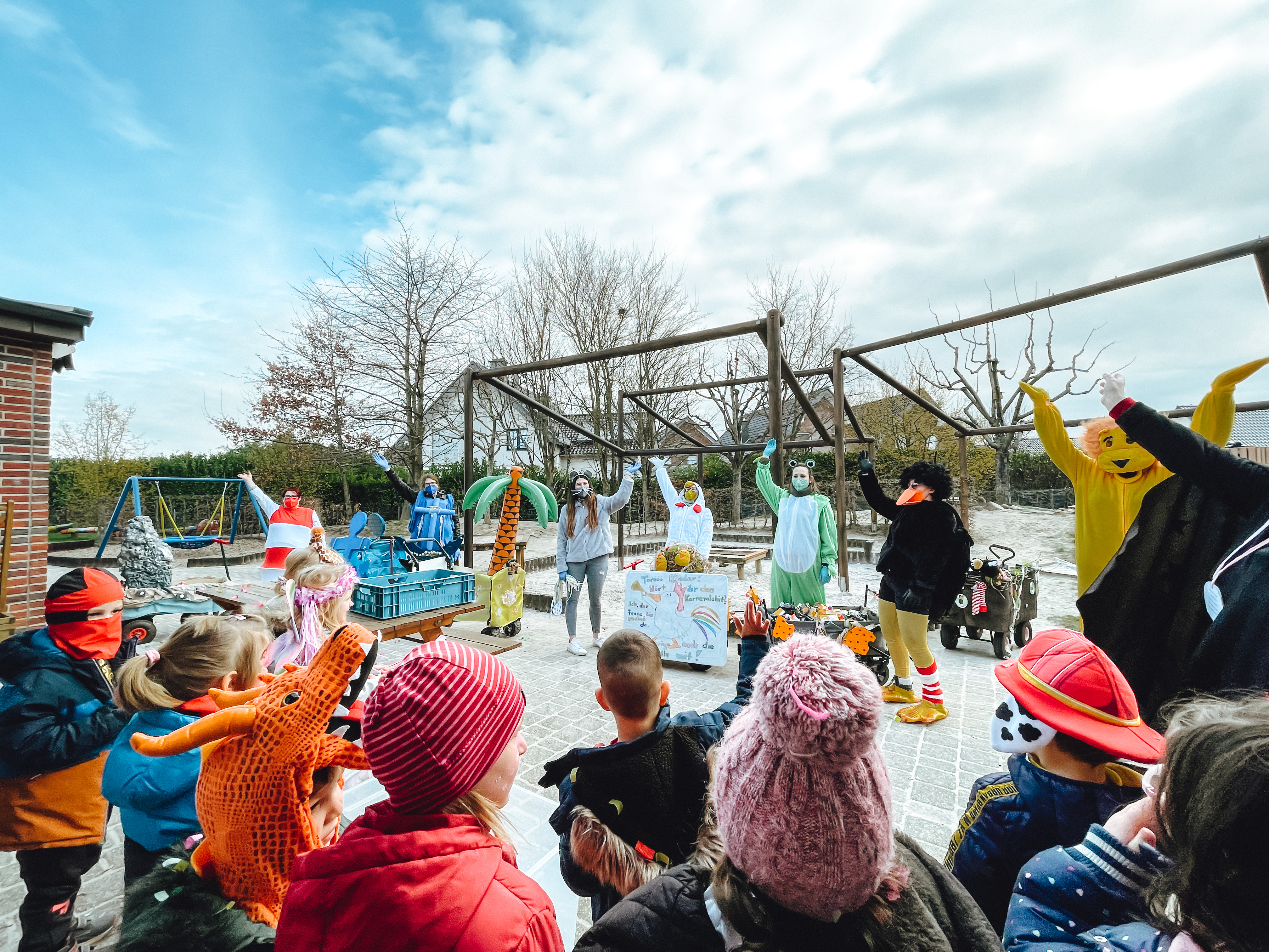 Kinder feiern draußen mit einem Maskottchen. Manche tragen Kostüme und Hüte. Es gibt einen kleinen See und einen Spielplatz im Hintergrund.