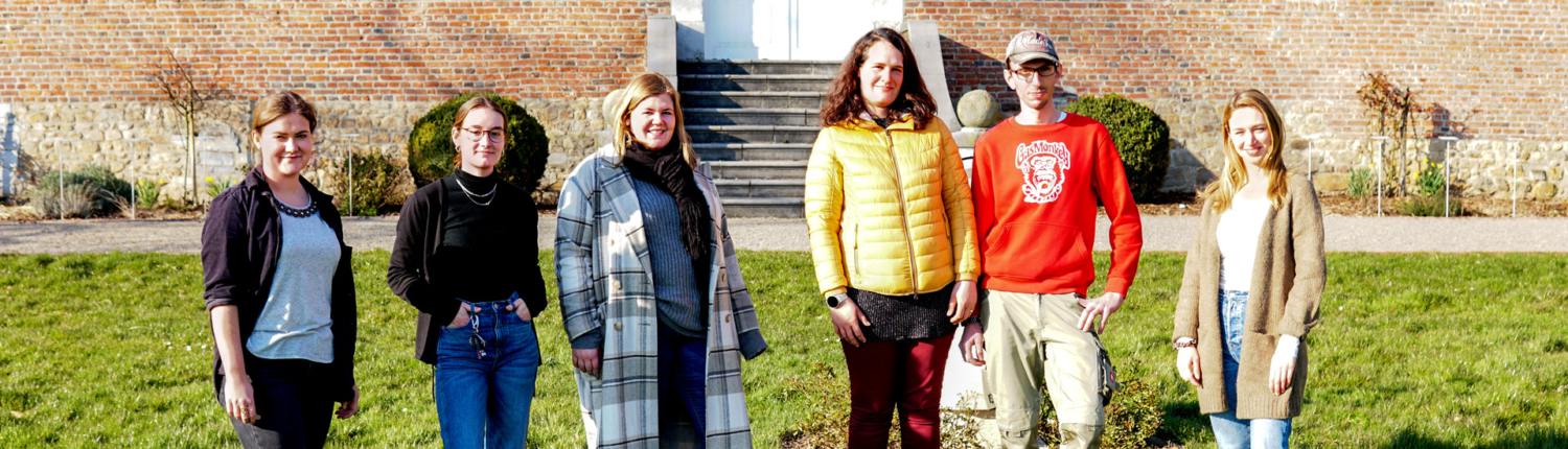 Gruppe von sechs Personen, die vor einem historischen Gebäude stehen, umgeben von Rasen und Blumen. Sie lächeln und tragen lässige Kleidung, während sie den sonnigen Tag genießen.