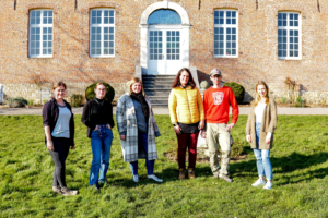 Gruppe von sechs Personen, die vor einem historischen Gebäude stehen, umgeben von Rasen und Blumen. Sie lächeln und tragen lässige Kleidung, während sie den sonnigen Tag genießen.