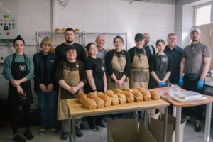 Bäckerteam in einer Bäckerei, posierend vor frisch gebackenem Brot auf einem Tisch. Die Mitarbeiter tragen Schürzen und lächeln in die Kamera, während die Bäckerei eine einladende Atmosphäre ausstrahlt.