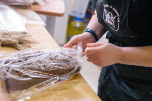 Eine Person wickelt ein frisch gebackenes Brot in Folie, um es frisch zu halten. Der Fokus liegt auf den Händen und dem Brot auf einer Holzoberfläche.