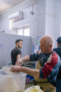 Ein Mann backt Brot. Er hat einen Kopfschleier und eine Schürze an. Er mischt etwas in einer Schüssel.