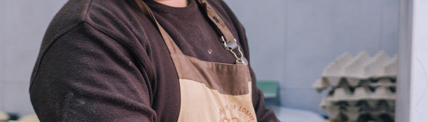 Lächelnder Bäcker in einer Backstube, der Teig vorbereitet und mit einem Holzbrett arbeitet. Im Hintergrund sind Backutensilien und Zutaten sichtbar.