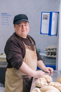 Ein Mann mit Schürze knetet Brot auf einem Tisch. Er trägt eine Mütze.