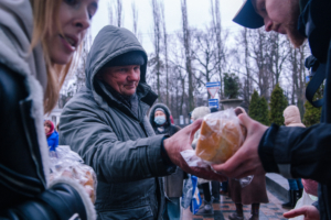 Menschen stehen an einer Essensausgabe und erhalten Lebensmittel aus Tüten. Die Szene zeigt eine kalte, graue Umgebung, in der Bedürftige Unterstützung erhalten.