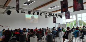 Teilnehmer bei einer Präsentation in einem modernen Veranstaltungsraum, mit einem großen Bildschirm im Hintergrund, der eine Szene zeigt. Die Zuschauer sitzen in Reihen und sind auf die Präsentation fokussiert.