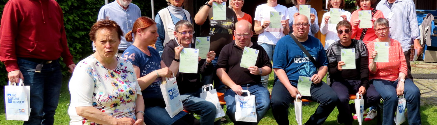 Gruppe von Menschen im Freien, die stolz ihre Urkunden oder Auszeichnungen halten, umgeben von grünem Gras und einem Gartenhaus, bei sonnigem Wetter.