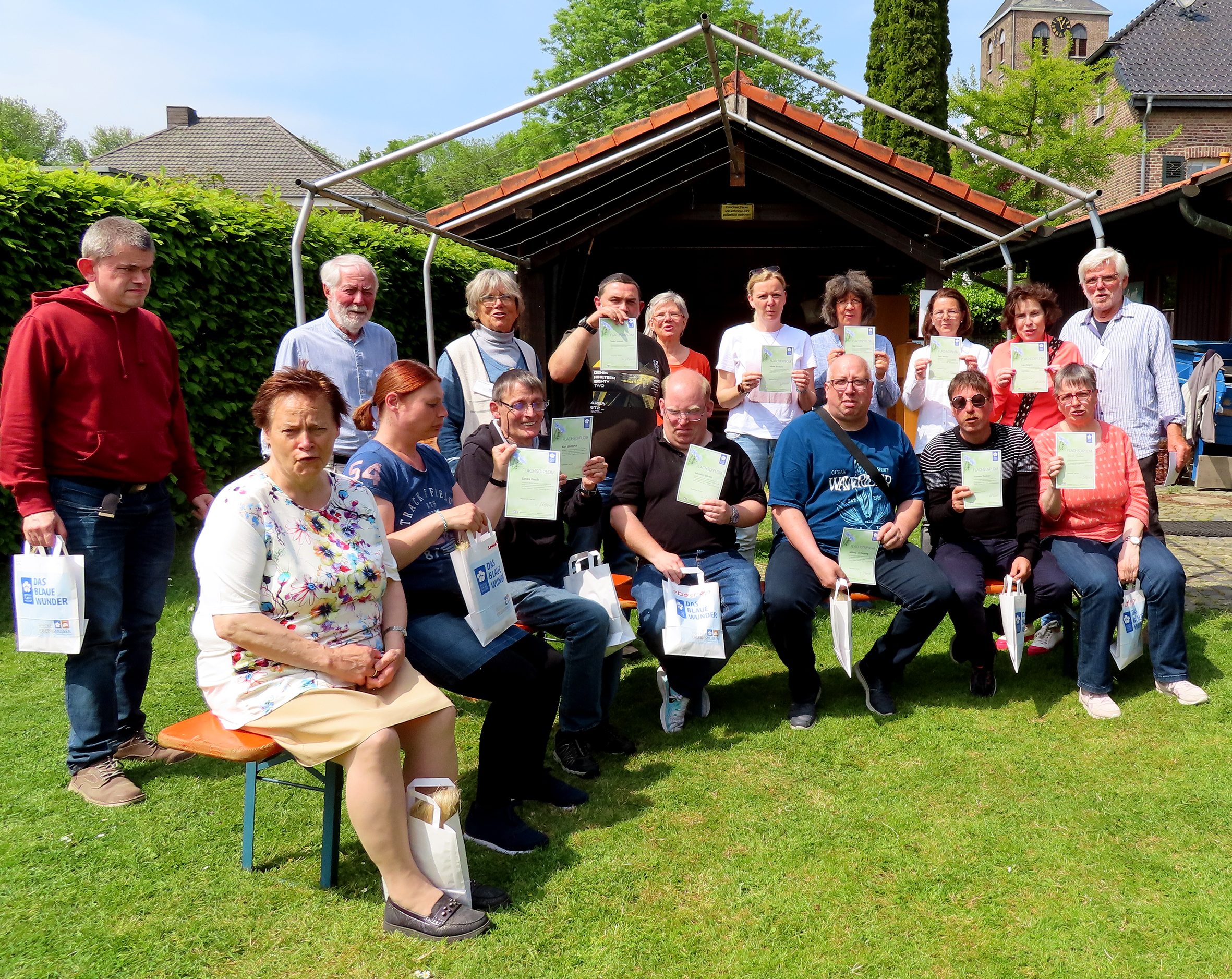 Gruppe von Menschen im Freien, die stolz ihre Urkunden oder Auszeichnungen halten, umgeben von grünem Gras und einem Gartenhaus, bei sonnigem Wetter.