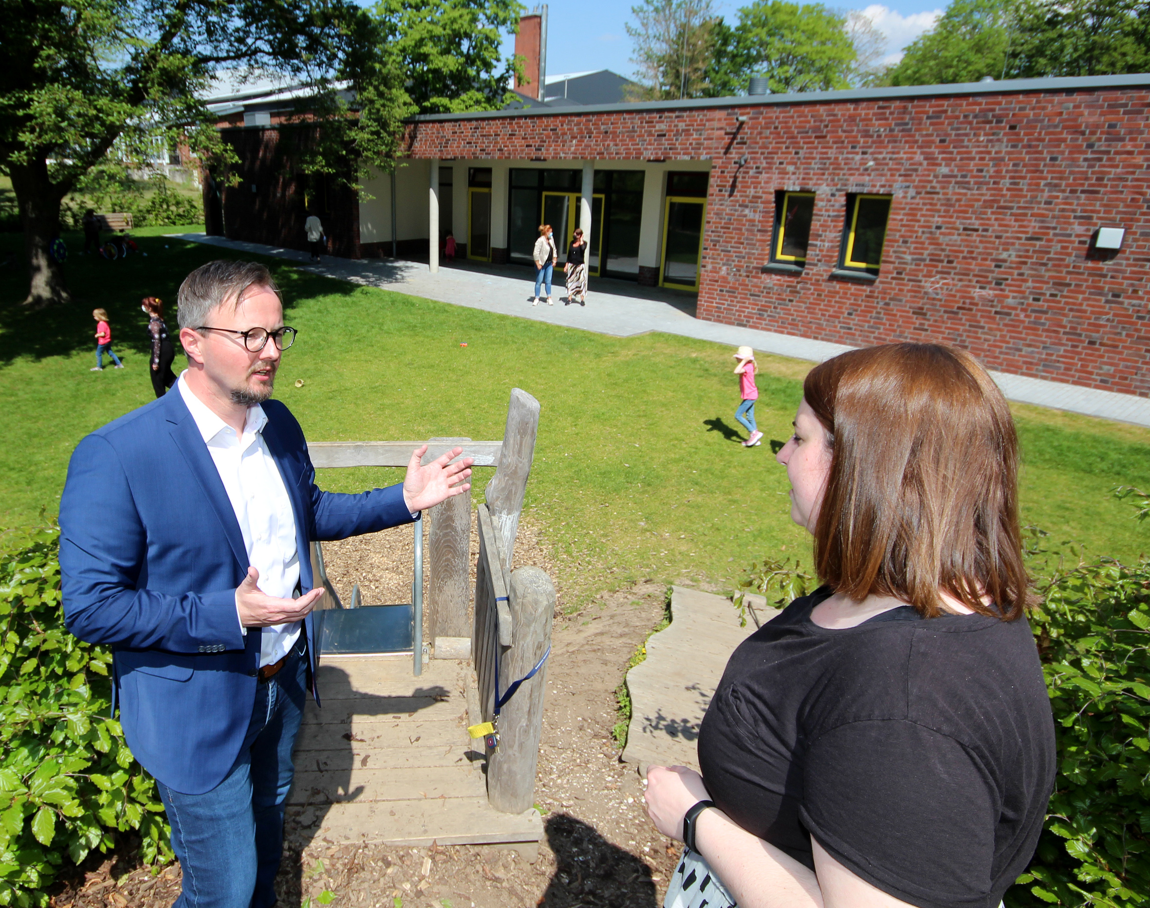 Mann im Anzug spricht mit einer Frau im Freien, umgeben von Pflanzen und einem modernen Gebäude im Hintergrund. Ideal für Themen wie Bildung, Kommunikation oder Gemeinschaftsprojekte.