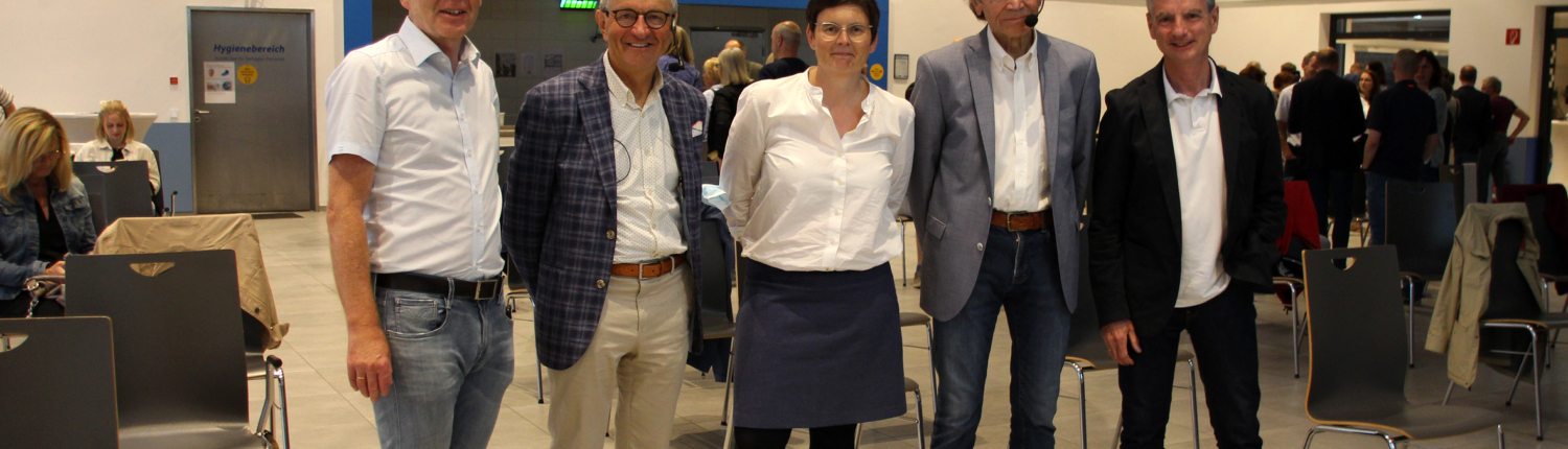 Gruppe von fünf Personen, die in einem Veranstaltungsraum stehen, während sie an einer Sitzung oder Versammlung teilnehmen. Alle sind formell gekleidet und lächeln in die Kamera.