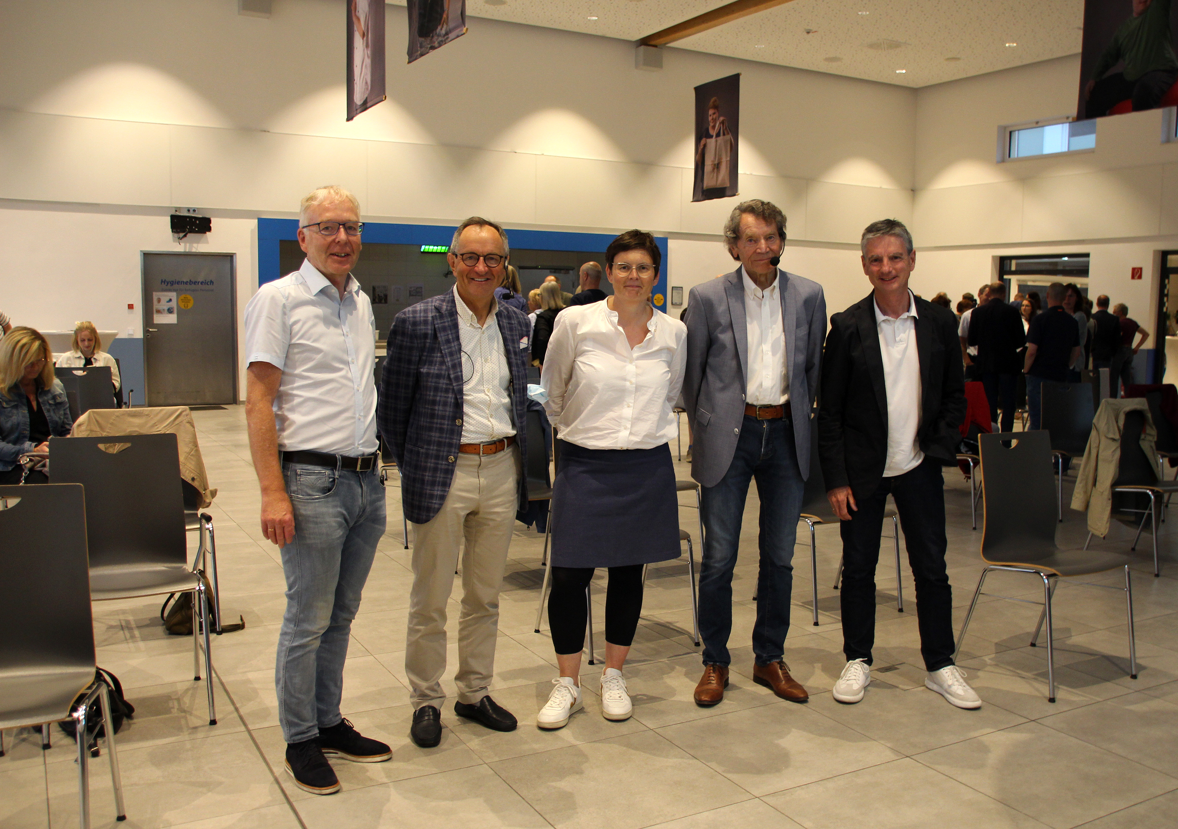 Gruppe von fünf Personen, die in einem Veranstaltungsraum stehen, während sie an einer Sitzung oder Versammlung teilnehmen. Alle sind formell gekleidet und lächeln in die Kamera.