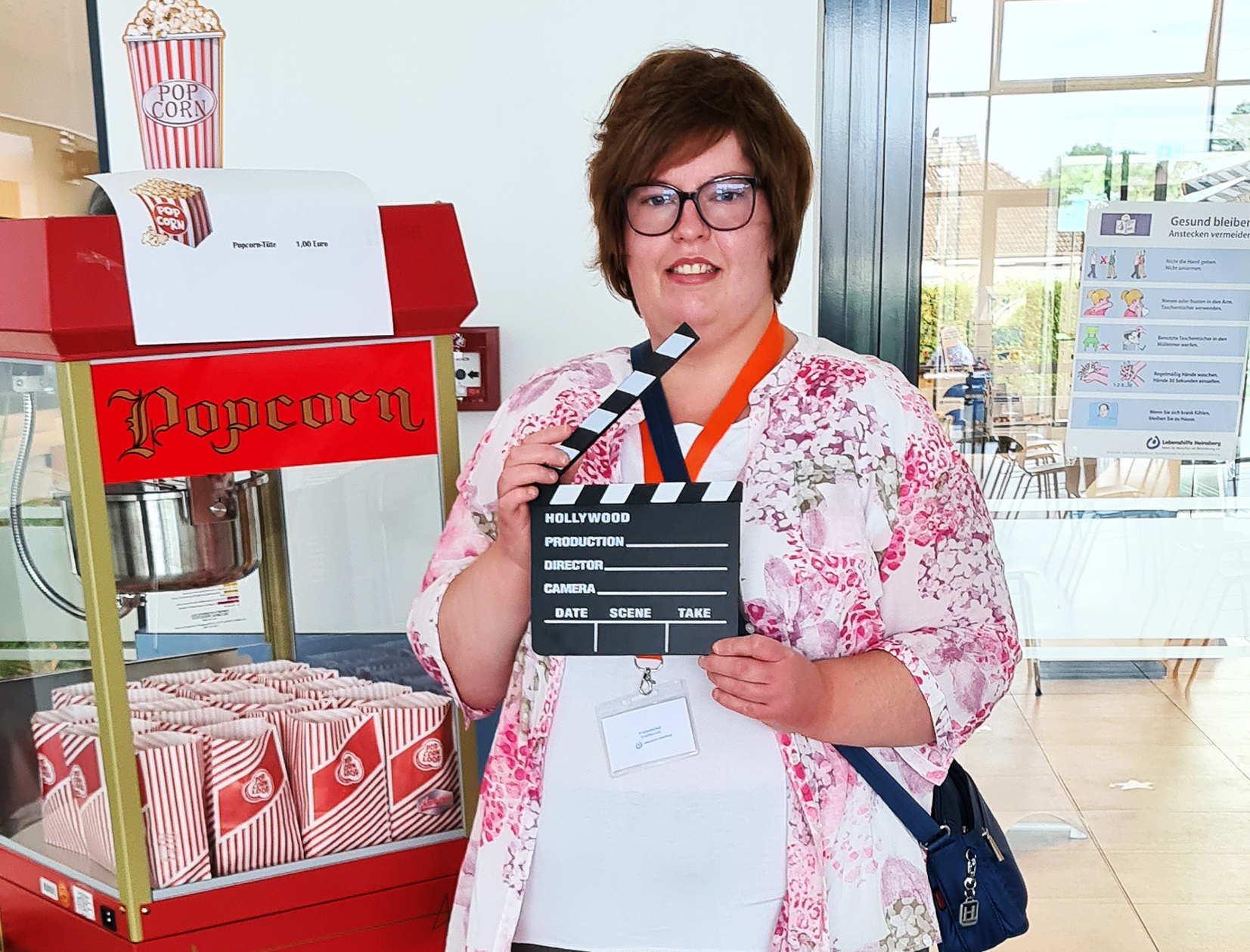 Frau mit Klappe und buntem Outfit steht vor einem Popcornstand auf einer Veranstaltung.