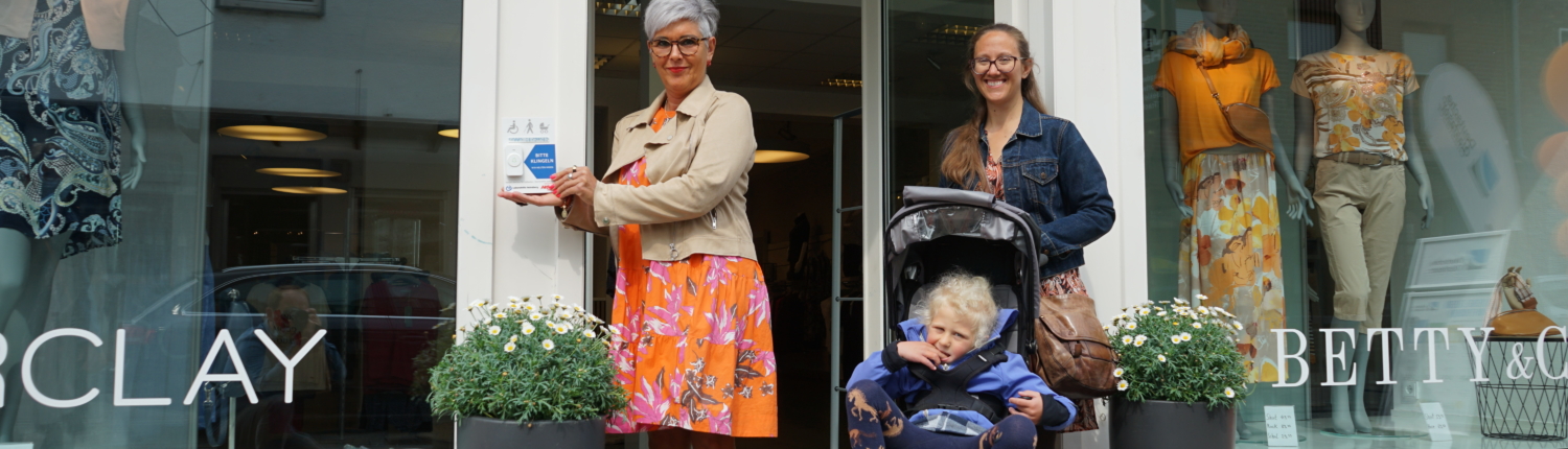 Zwei Frauen stehen an der Eingangstür eines Geschäfts, eine von ihnen hält ein blaues Band, während die andere einen Kinderwagen schiebt. Die Szene vermittelt eine feierliche Eröffnung oder einen besonderen Anlass.