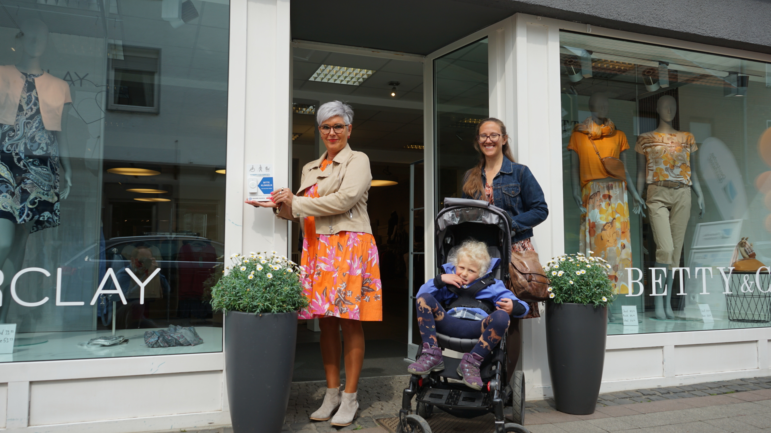 Zwei Frauen stehen an der Eingangstür eines Geschäfts, eine von ihnen hält ein blaues Band, während die andere einen Kinderwagen schiebt. Die Szene vermittelt eine feierliche Eröffnung oder einen besonderen Anlass.