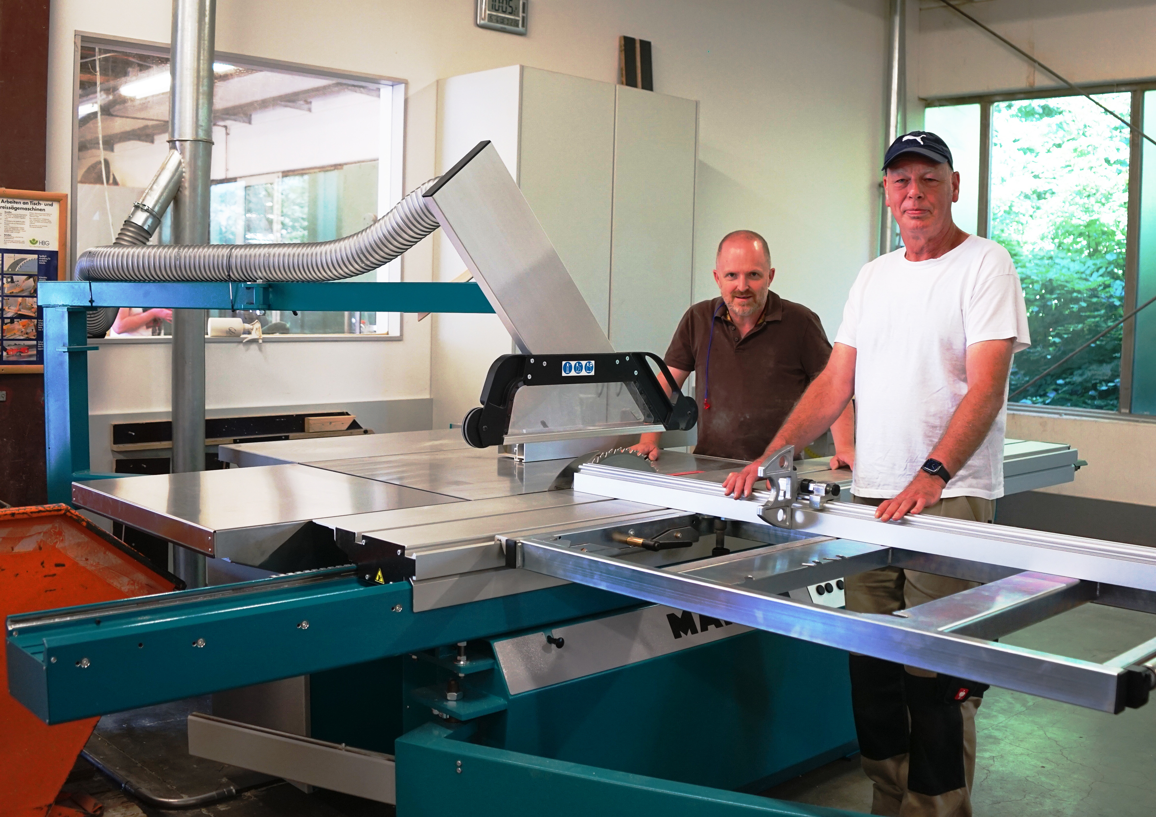 Zwei Männer stehen vor einer großen Säge. Einer hält ein Werkzeug.