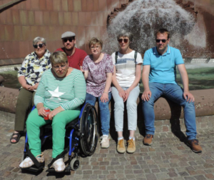 Fünf Personen sitzen vor einem Springbrunnen. Zwei tragen Hüte, drei tragen T-Shirts. Eine Person sitzt im Rollstuhl.