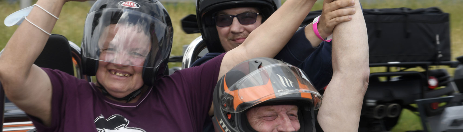 Person sitzt auf einem Motorrad. Die Person trägt einen Helm und eine Brille. Im Hintergrund sind Bäume und ein Auto zu sehen.