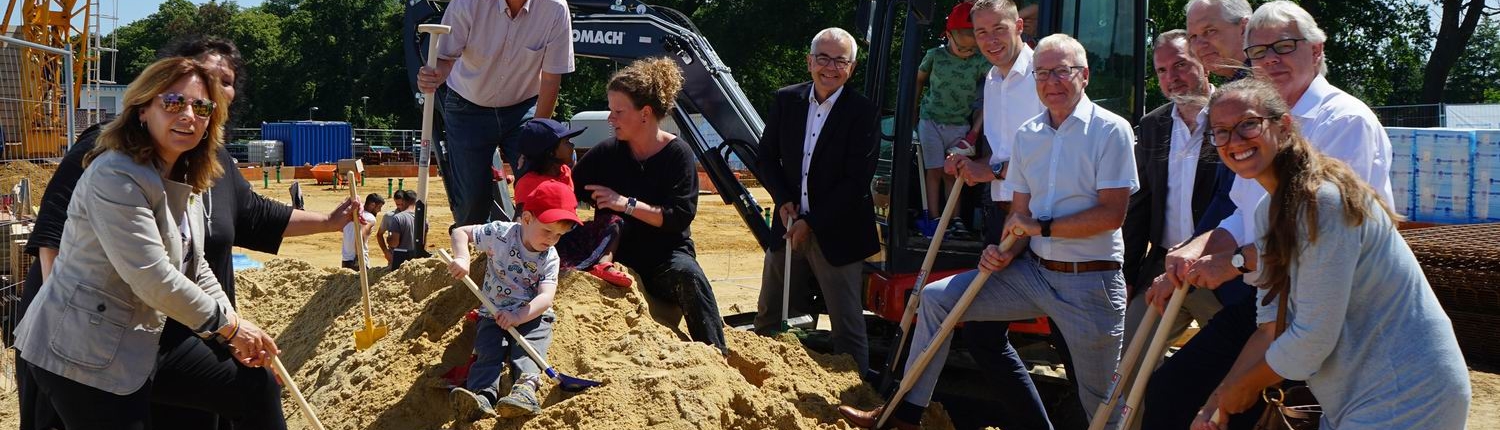 Feierliche Grundsteinlegung mit mehreren Personen, darunter Kinder, die mit Schaufeln im Sand spielen, vor einem Bagger und einer Baustelle im Freien.