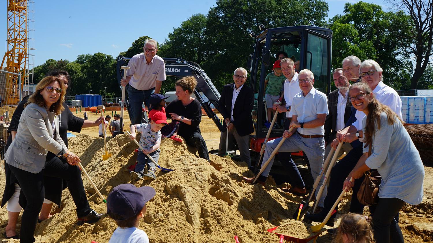 Feierliche Grundsteinlegung mit mehreren Personen, darunter Kinder, die mit Schaufeln im Sand spielen, vor einem Bagger und einer Baustelle im Freien.