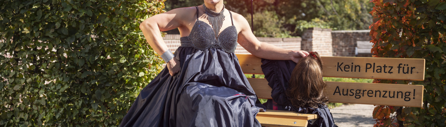 Frau in einem eleganten, dunkelblauen Abendkleid sitzt auf einer Bank in einem Garten. Im Hintergrund sind grüne Sträucher und ein rotes Licht sichtbar. Die Szene strahlt eine kreative und künstlerische Atmosphäre aus.