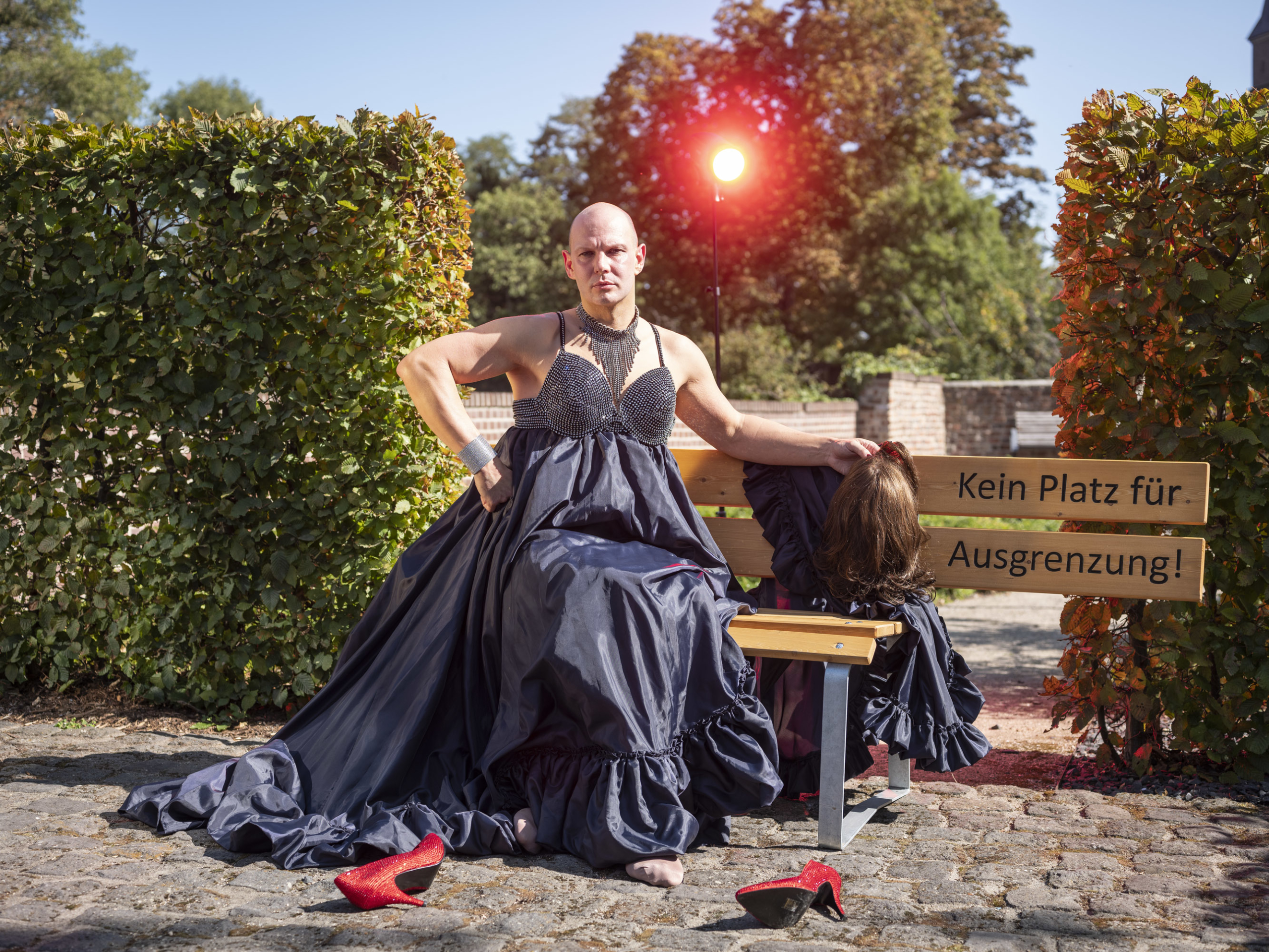 Frau in einem eleganten, dunkelblauen Abendkleid sitzt auf einer Bank in einem Garten. Im Hintergrund sind grüne Sträucher und ein rotes Licht sichtbar. Die Szene strahlt eine kreative und künstlerische Atmosphäre aus.