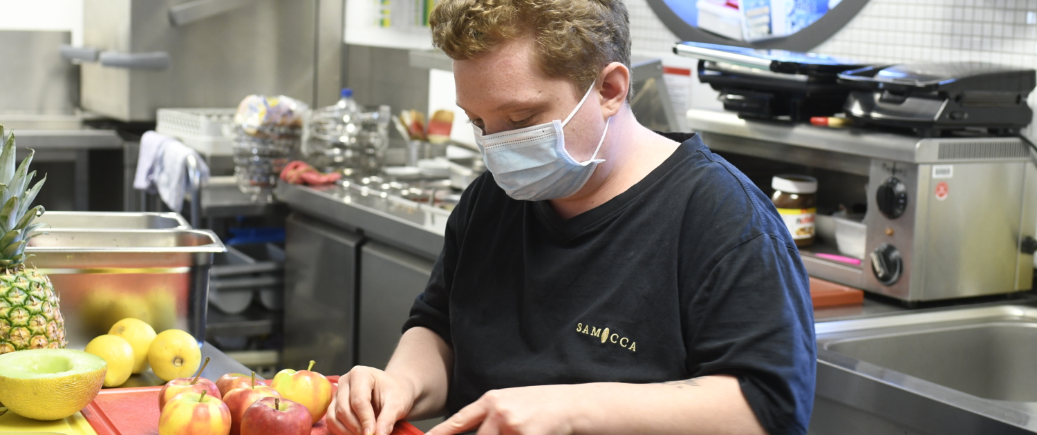 Koch in einer modernen Küche schneidet Obst auf einem roten Schneidebrett, während er eine Maske trägt.