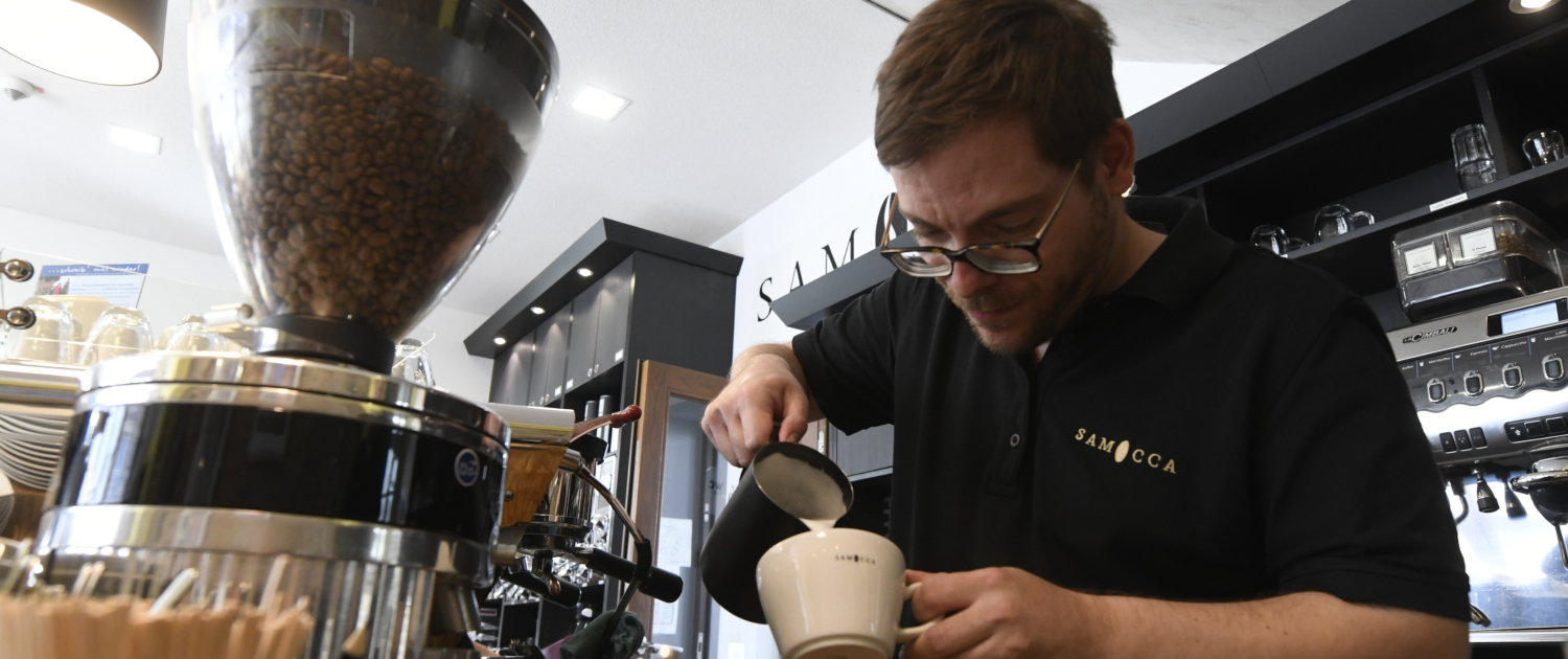 Ein Mann gießt Kaffee um. Er steht in einem Café und benutzt eine Kaffeemaschine.